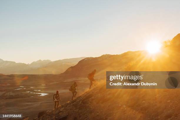 a group of hikers with backpacks walking uphill at sunset to the top of the mountain. travel and adventure concept - colina acima imagens e fotografias de stock