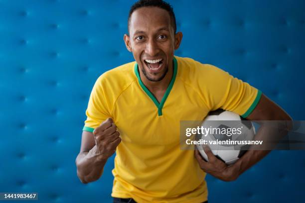 portrait of brazilian man with brazilian soccer team shirt, holding soccer ball in front of blue background - intercontinental trophy stock pictures, royalty-free photos & images