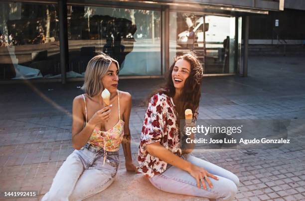 two young girls talk and have fun and eat ice cream sitting in their city park on a summer day - hot spanish women ストックフォトと画像