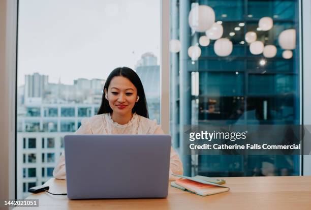 A young woman sits at a table in an office environment and uses a laptop