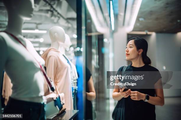 beautiful young asian woman checking her smartphone while standing outside a boutique looking at window display in shopping mall. window shopping. sale season - window shopping stock pictures, royalty-free photos & images