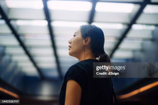 confident young asian businesswoman looking up with optimistic expression, standing in a building with opened roof top in the city. planning for the future. feeling hopeful and bright. strive for success and achievement - energy independence stock pictures, royalty-free photos & images