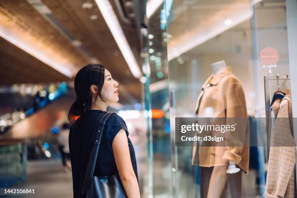 young asian woman standing outside a boutique looking at window display while shopping in shopping mall. window shopping. sale season - luxus shopping stock-fotos und bilder