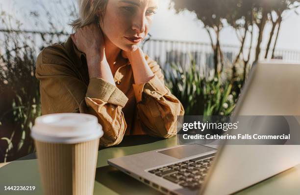 a woman sits at a laptop, holding her neck. she looks stressed. - parte del cuerpo humano fotos fotografías e imágenes de stock