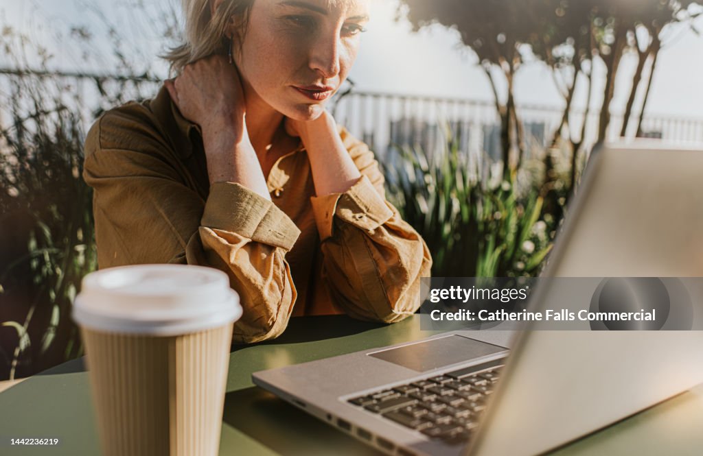 A woman sits at a laptop, holding her neck. She looks stressed.
