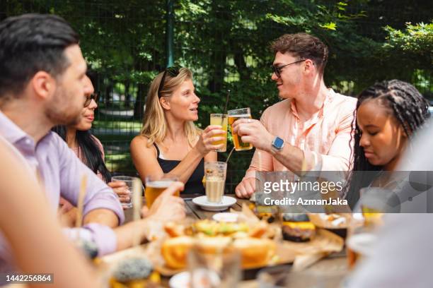 mujer caucásica y hombre caucásico brindando con bebidas frescas en una reunión de amigos afuera - male burger eating fotografías e imágenes de stock