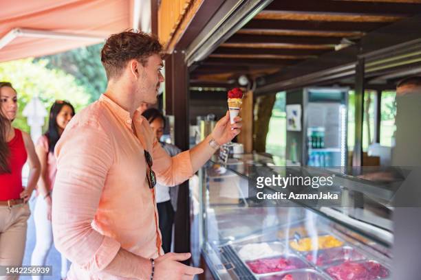 junger attraktiver mann, der eine eistüte mit zwei geschmacksrichtungen von der theke einer eisdiele holt - ice cream counter stock-fotos und bilder