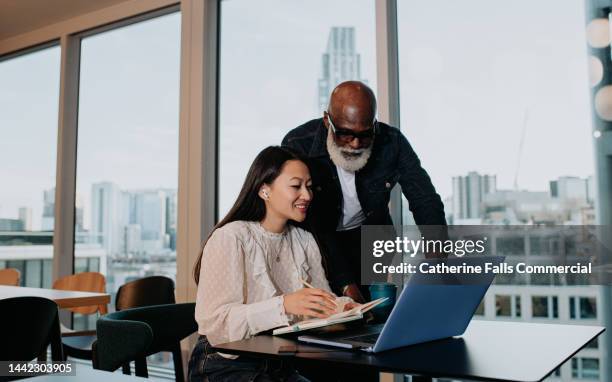 a older man stands behind a young woman and looks over her shoulder at her laptop - boss over shoulder stock-fotos und bilder