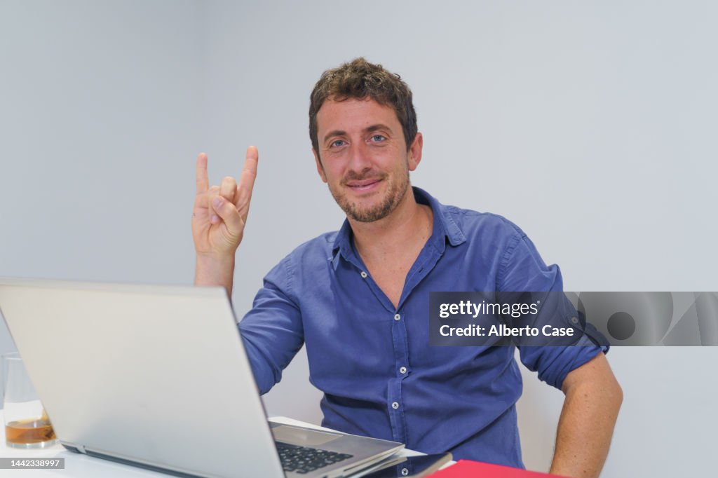 Happy businessman looking at camera and making heavy metal hand sign in office