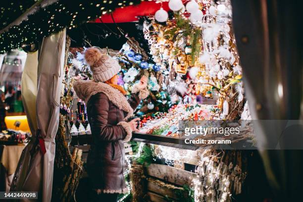 frau in warmen kleidern blickt auf beleuchteten weihnachtsmarktstand - bazaar market stock-fotos und bilder