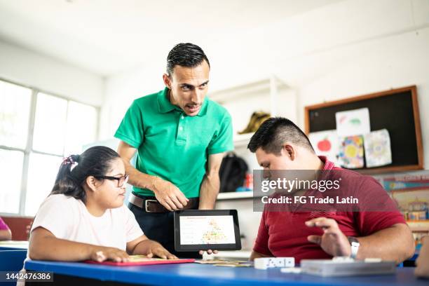 mid adult teacher with a cognitive disability showing a lesson on a digital tablet to two students in the classroom - developmental disability stock pictures, royalty-free photos & images