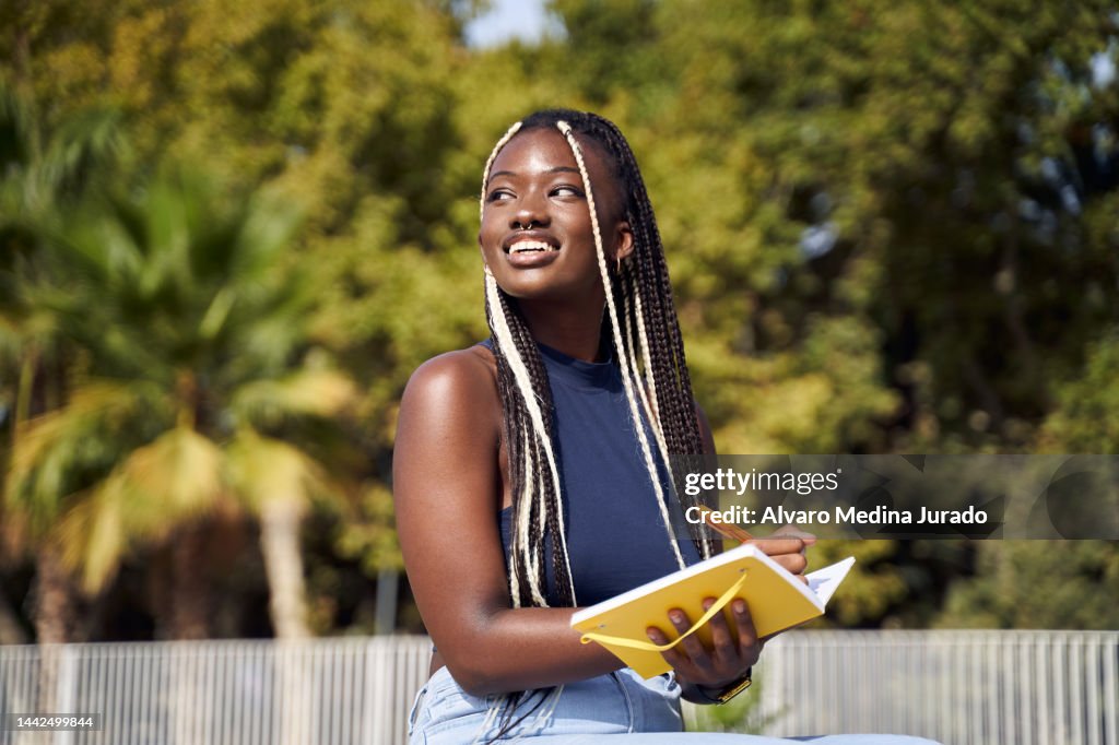 Cheerful young African American girl outdoors in the city reading and writing in a notebook while sitting in a park.