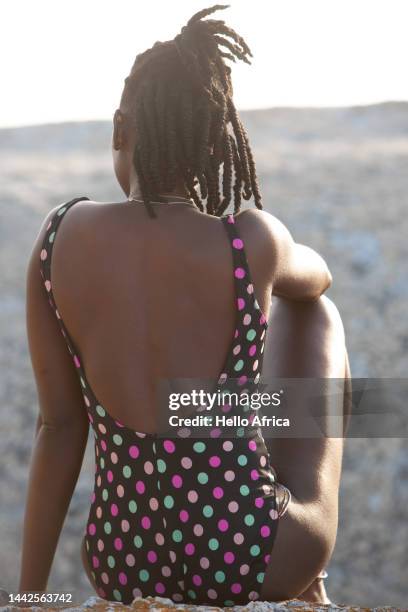 a beautiful young woman sits with her back to camera, a twenty something wearing a black polka dot swimming costume with colourful spots & with an afro hairstyle tied in natural braids looks away with a natural clear blue sky as background - female looking away from camera serious thinking outside natural stock-fotos und bilder