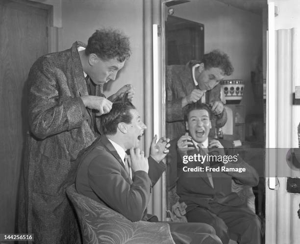 English actor and comedian Frankie Howerd cuts the hair of entertainer Max Bygraves in their shared dressing room during rehearsals for the Royal...