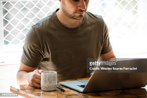 man with green t-shirt typing on a laptop on the balcony and holding a cup of coffee - caffeine molecule stock pictures, royalty-free photos & images