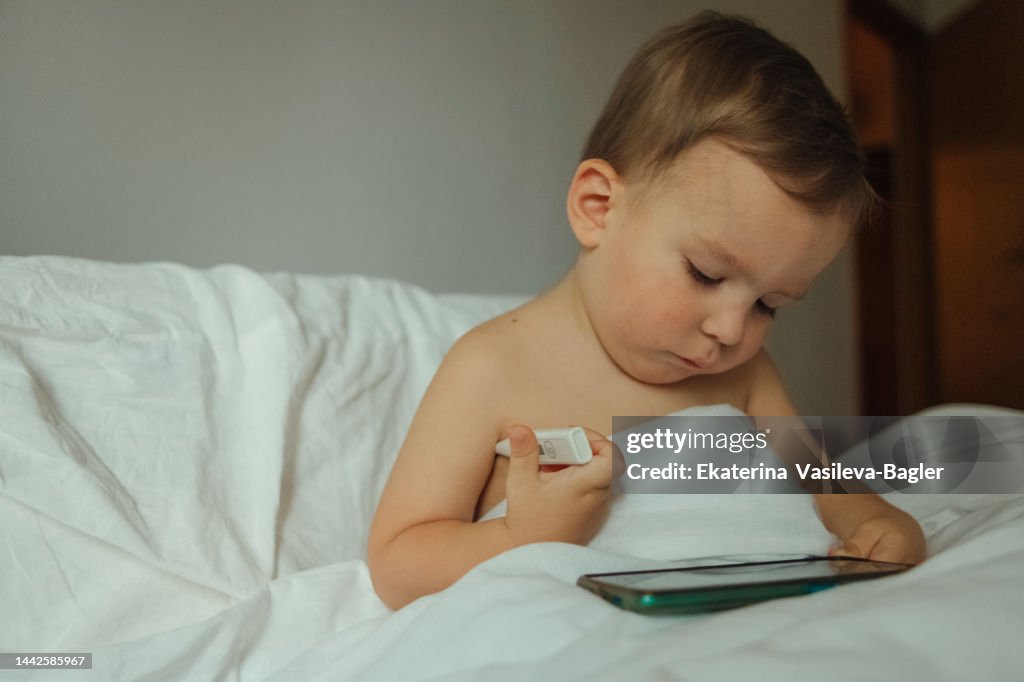 A child 2-3 years old sits on the bed measures the temperature with an electronic thermometer and looks at the mobile phone