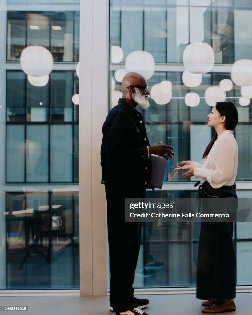Co-workers stand face-to-face beside a huge window and have a conversation
