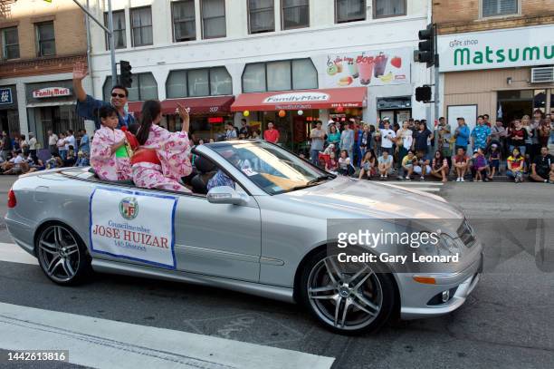 At the Little Tokyo Nisei Week Parade Council Member José Huizar rides in a silver Mercedes convertible with two girls in kimonos and waves smiling...