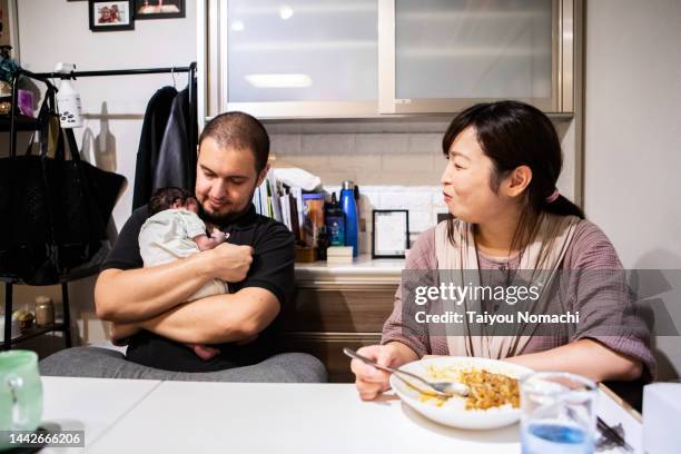 a father holding his sleeping newborn daughter in his arms and a mother smiling and eating curry next to him. - yōshoku stockfoto's en -beelden