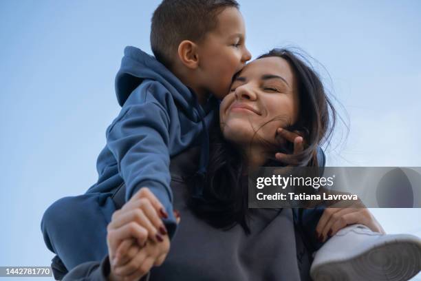 happy small boy kissing his mother. smiling woman giving piggyback ride to a small boy outside. lifestyle happy family concept - happy mothers day mom stock-fotos und bilder