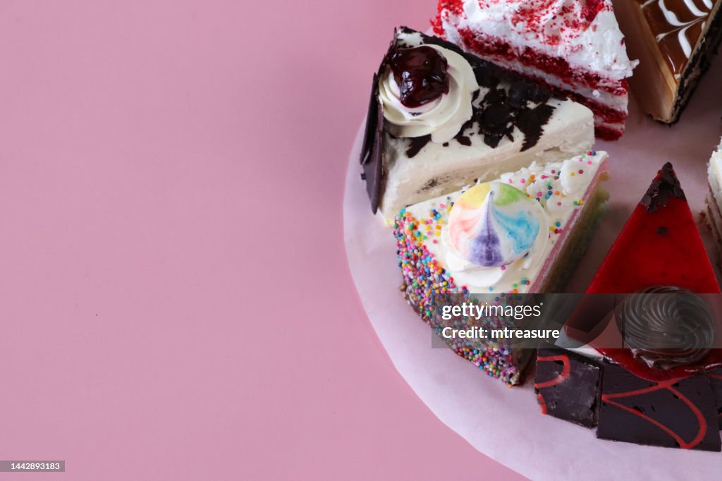 Close-up image of slices of gateaux varieties on circle of greaseproof parchment paper, chocolate cherry cake, Black Forest gateau, red velvet, rainbow cake, coffee marbled chocolate gateau, pink background, focus on foreground, elevated view, copy space