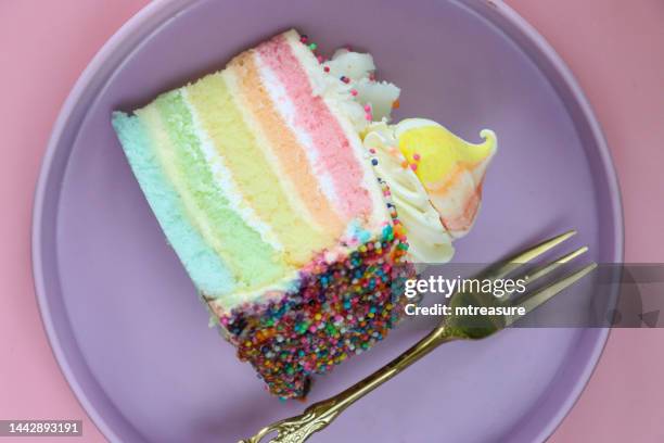 immagine ravvicinata della fetta di torta arcobaleno, cinque strati di pan di spagna arcobaleno separati da crema al burro alla vaniglia su piatto viola con forchetta, rosetta di glassa in filo, bacio di meringa arcobaleno, sfondo rosa, vista elevata - gateaux foto e immagini stock