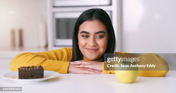 woman choosing between a cake or apple in the kitchen to eat as a day time snack at home. indian girl thinking about the healthy, nutrition and organic fruit or the unhealthy chocolate dessert. - healthy indulgence stock pictures, royalty-free photos & images