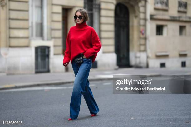 Diane Batoukina wears black sunglasses, a red wool ribbed turtleneck pullover, a black shiny leather belt bag from Chanel, blue faded denim wide legs...