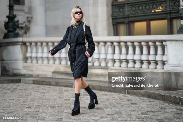 Natalia Verza wears black futurist sunglasses from Chanel, gold earrings, a black hoodie long parka rain coat, a white shiny leather with gold jewels...
