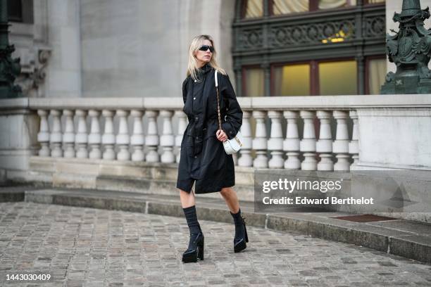 Natalia Verza wears black futurist sunglasses from Chanel, gold earrings, a black hoodie long parka rain coat, a white shiny leather with gold jewels...