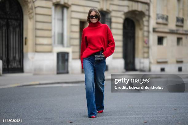 Diane Batoukina wears black sunglasses, a red wool ribbed turtleneck pullover, a black shiny leather belt bag from Chanel, blue faded denim wide legs...