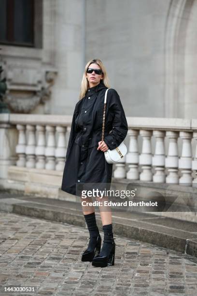 Natalia Verza wears black futurist sunglasses from Chanel, gold earrings, a black hoodie long parka rain coat, a white shiny leather with gold jewels...