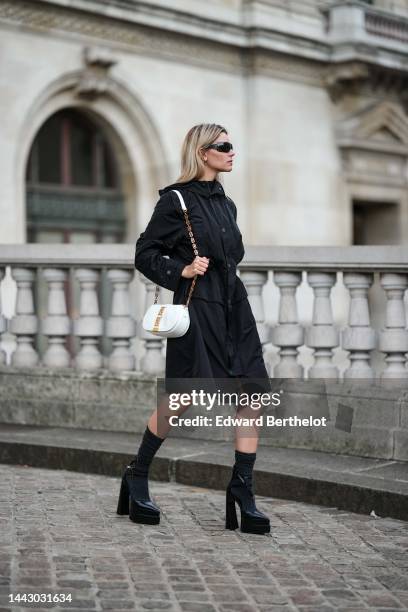 Natalia Verza wears black futurist sunglasses from Chanel, gold earrings, a black hoodie long parka rain coat, a white shiny leather with gold jewels...