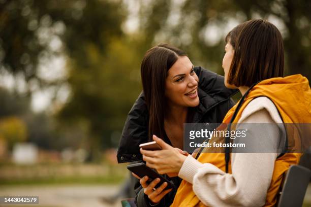 two girlfriends sitting on the street bench, looking at social media posts via smart phone, chatting and bonding - girlfriend meme stock pictures, royalty-free photos & images