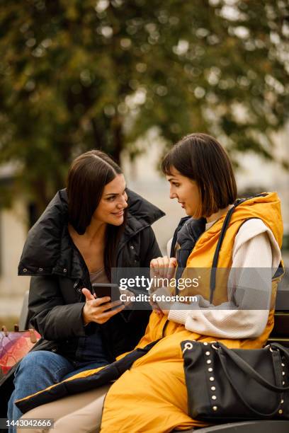 two girlfriends sitting on the street bench, looking at social media posts via smart phone, chatting and bonding - girlfriend meme stock pictures, royalty-free photos & images