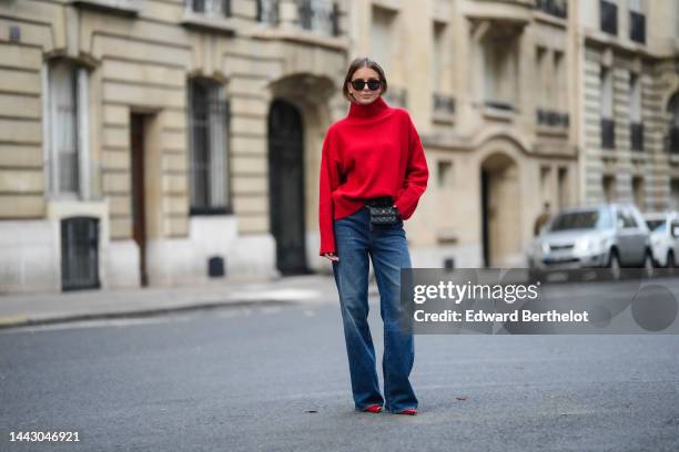 Diane Batoukina wears black sunglasses, a red wool ribbed turtleneck pullover, a black shiny leather belt bag from Chanel, blue faded denim wide legs...