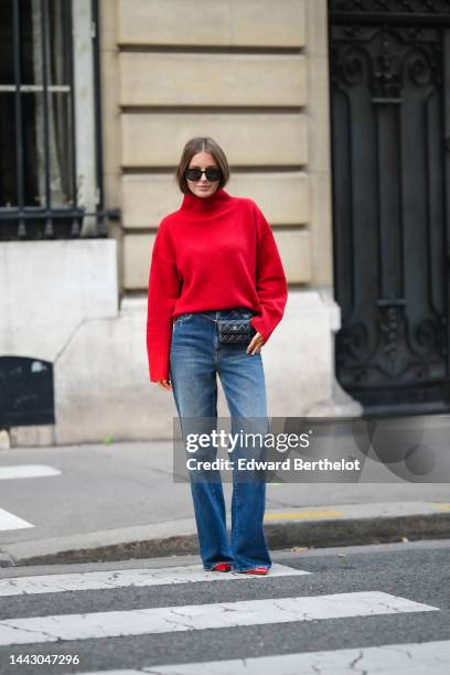 Diane Batoukina wears black sunglasses, a red wool ribbed turtleneck pullover, a black shiny leather belt bag from Chanel, blue faded denim wide legs...