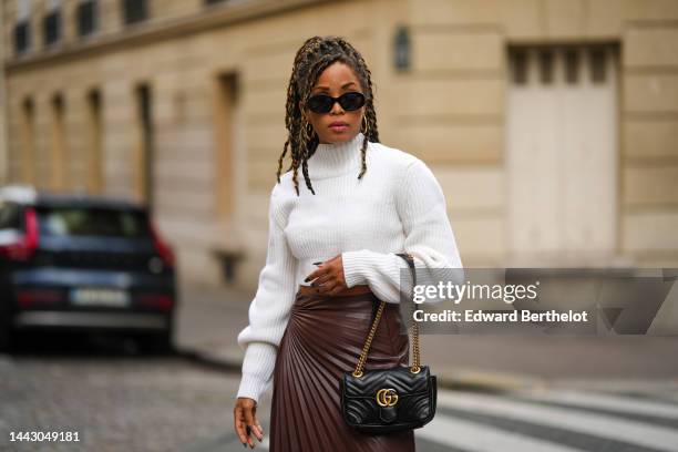 Ellie Delphine wears black sunglasses from Celine, gold earrings, a white ribbed wool high neck / puffy long sleeves / cropped top, a black shiny...