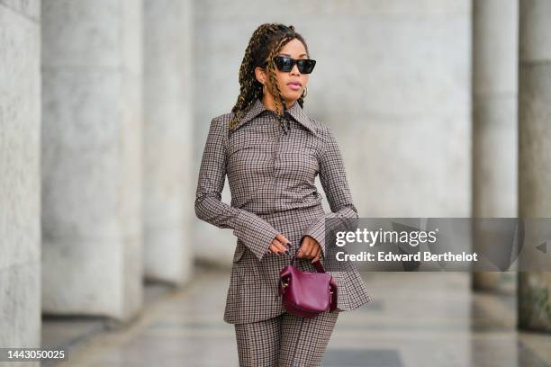 Ellie Delphine wears black cat eyes sunglasses, gold earrings, a black and brown checkered print pattern shirt, a matching black and brown checkered...