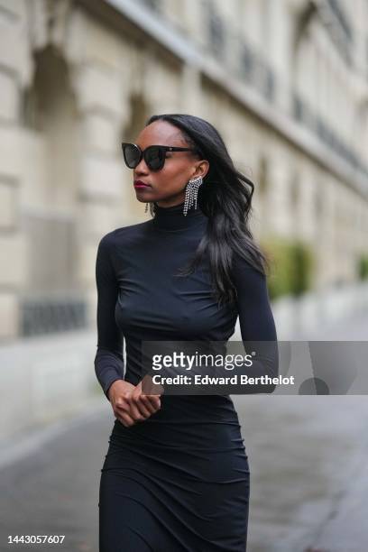 Emilie Joseph wears black sunglasses, a silver and rhinestones fringed pendant earrings, a black turtleneck / long sleeves / backless / long tube...