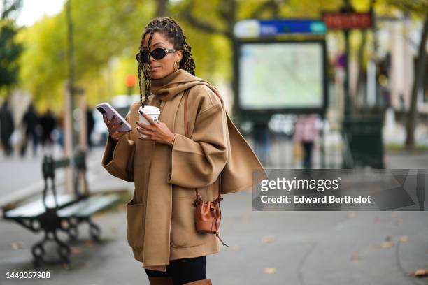 Ellie Delphine wears black sunglasses from Celine, gold earrings, a beige long wool scarf, a matching beige wool buttoned coat, black legging pants,...
