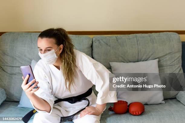 blonde girl with a mask practicing karate at home. resting on the couch after training. looking at her cell phone. black karate belt and red gloves. quarantine virus. home isolated. - karate girl isolated stock pictures, royalty-free photos & images