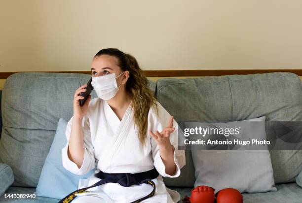 blonde girl with a mask practicing karate at home. resting on the couch after training. talking on her cell phone. black karate belt and red gloves. quarantine virus. home isolated. - karate girl isolated stock pictures, royalty-free photos & images