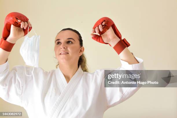 blonde girl in a mask practicing karate at home. taking off her mask. symbol of victory. karate black belt and red gloves. quarantine virus. home isolated. - karate girl isolated stock pictures, royalty-free photos & images