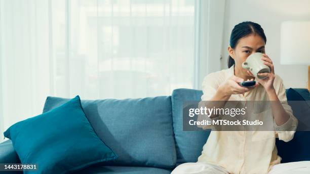 beautiful asian young woman on sofa drink coffee click remote controller change channel on television choose favorite movie in streaming online vod, video on demand at home. - pleased face laptop stock pictures, royalty-free photos & images