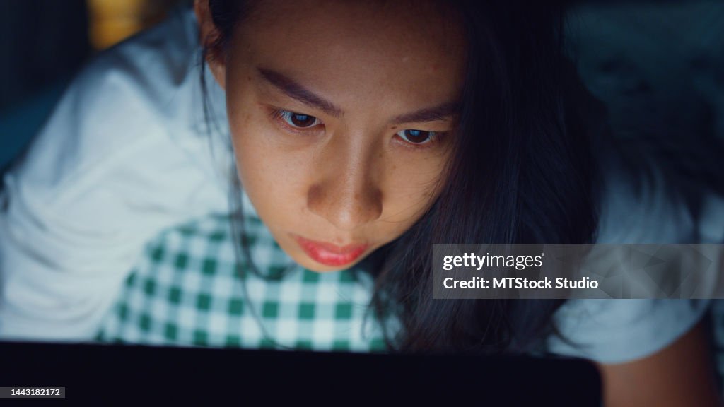 Exhausted Young Asian girl laying on bed feel stress eyes focus on computer laptop screen working late overtime in bedroom at night.