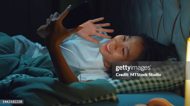 close-up young asia girl media addiction laying on bed wear earphone use cell phone video call with boyfriend before bedtime on in bedroom at night. - lange afstandsrelatie stockfoto's en -beelden