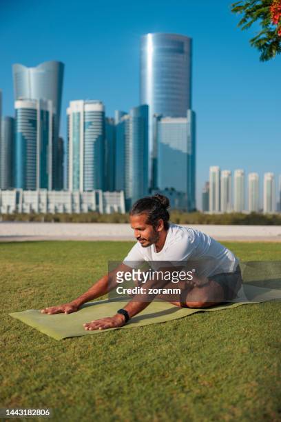 bärtiger mann praktiziert yoga auf dem rasen des öffentlichen parks in abu dhabi, sitzt im schneidersitz und beugt sich nach vorne - holy city park stock-fotos und bilder