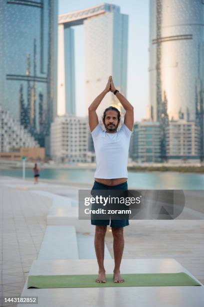 bearded man standing on a mat and exercising outdoors, clasped hands above head - yoga office arab stock pictures, royalty-free photos & images