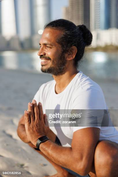 bearded man with hair bun doing yoga on sandy beach in abu dhabi, smiling with hands clasped on chest - yoga office arab stock pictures, royalty-free photos & images